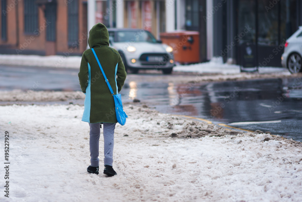 woman in green coat with blue bag walks along side road with traffic snow and slush in background