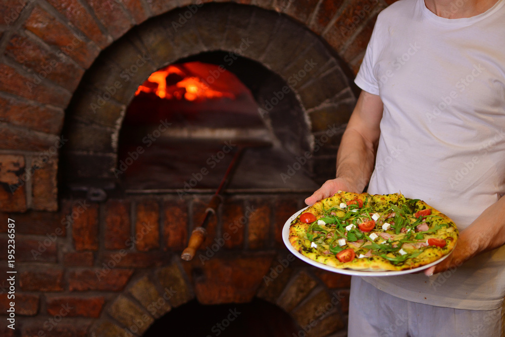 Chef holds a tasty pizza in his hands. Cook with a finished pizza in front of a wood stove.