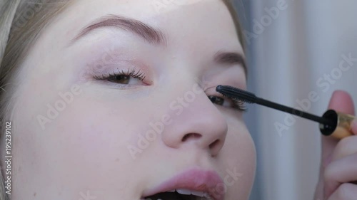 Beautiful blonde woman doing makeup in front of mirror. Smiling woman in pink dress behind mirror. Attractive girl doing makeup. Fashion and cosmetics concept, lips and eyes makeup.  