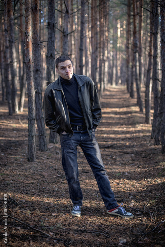 A young man of criminal appearance in a black leather jacket posing in an autumn forest.