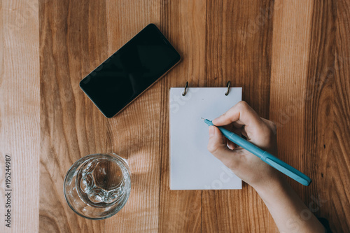 Business and office concept. Girl writing in notepad. Phone and glass. On wooden background. photo