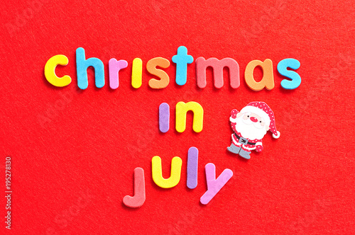The words christmas in July in colorful letters on a red background and a Santa Clause figurine