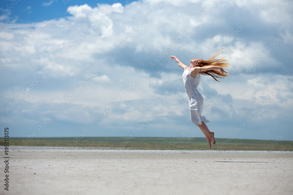 A woman in a white dress jumps high against a background of a sky landscape with a desert.
