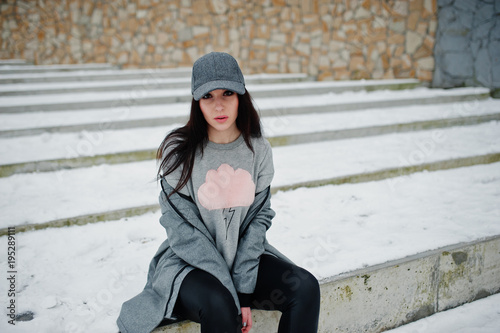 Stylish brunette girl in gray cap, casual street style on winter day against colored wall.
