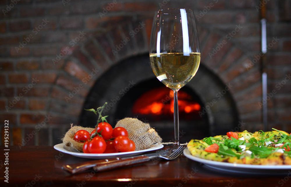 Wine in a wineglass with a pizza and cherry tomatoes near the old fireplace in a authentic restaurant.
