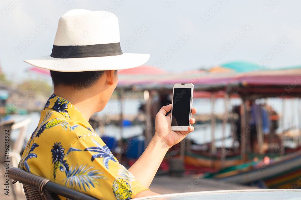 Fototapeta premium View from inside of young man wearing vintage style shirt with brim hat and using smartphone for communication at harbor. Travel in Chantaburi Province, Thailand. Soft focus and blur. Tourist concept.