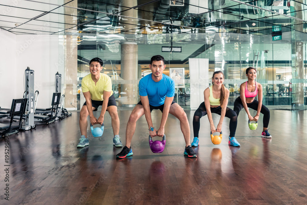 Full length view of four fit and cheerful young people smiling while holding heavy kettlebells during group class of functional training at the gym