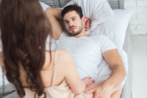 partial view of young man looking at girlfriend in pajamas on bed