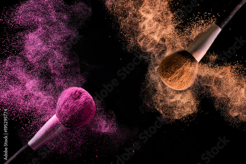  Two brushes for makeup with orange and pink make-up shadows in motion on a black background.