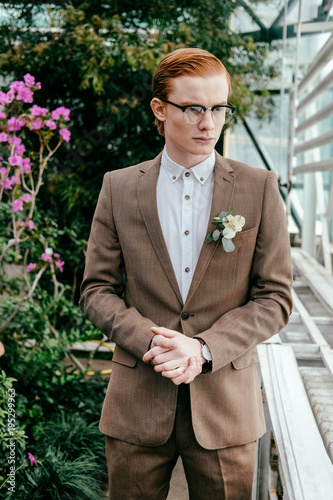 portrait of stylish man with red hair in suit and eyeglasses looking away in greenhouse