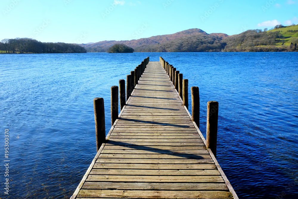 very long wooden symetrical beautiful wooden jetty, jutting out from the centre of the image into a calm blue lake with hills of forest and meadows in background