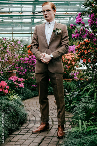 fashionable young man with red hair in suit and eyeglasses standing in glasshouse
