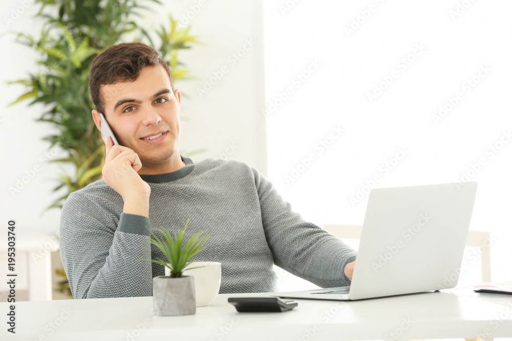 Young man talking on phone while working with laptop in home office Young man talking on phone while working with laptop in home office