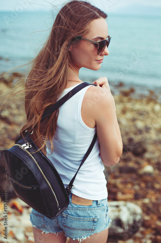 side view of young woman in sunglasses with backpack with ocean on background