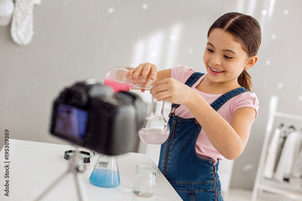 Little prodigy. Adorable pretty pre-teen girl pouring a pink chemical into the flask with water and smiling cheerfully while recording a video blog Little prodigy. Adorable pretty pre-teen girl pouring a pink chemical into the flask with water and smiling cheerfully while recording a video blog