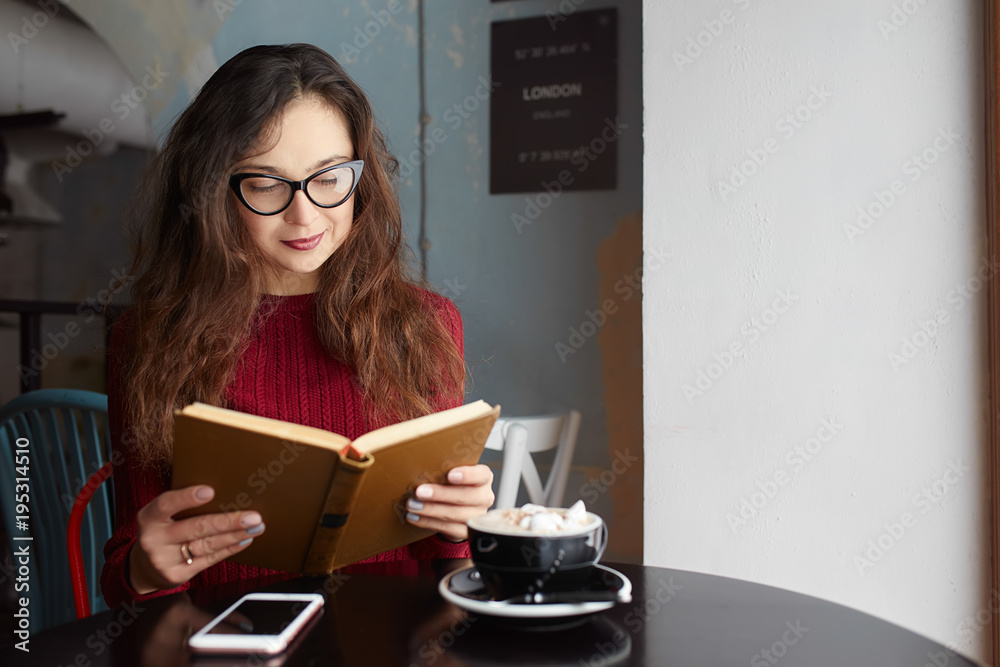 Young Beautiful Woman Reading Old Books, Using her Phone and Drinking Coffee or Cacao with Marshmallow. Relaxing atmosphere and Education. Young Beautiful Woman Reading Old Books, Using her Phone and Drinking Coffee or Cacao with Marshmallow. Relaxing atmosphere and Education.