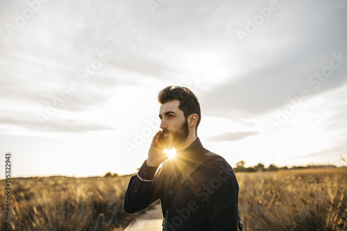 Content adult man with beard in stylish outfit speaking on phone against countryside in sunlight. © photopitu