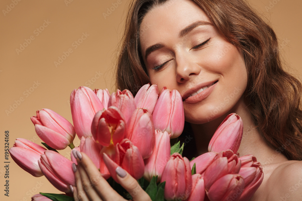 Closeup photo of gentle girl 20s with natural makeup smiling and holding lots of pink flowers near face, isolated over beige background Closeup photo of gentle girl 20s with natural makeup smiling and holding lots of pink flowers near face, isolated over beige background