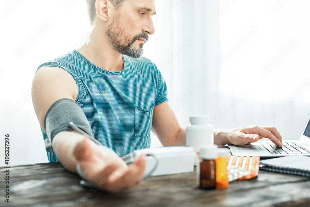Bad news. Tired upset unshaken man sitting at home in the bright room by the table looking at the laptop and measuring pulse. Bad news. Tired upset unshaken man sitting at home in the bright room by the table looking at the laptop and measuring pulse.