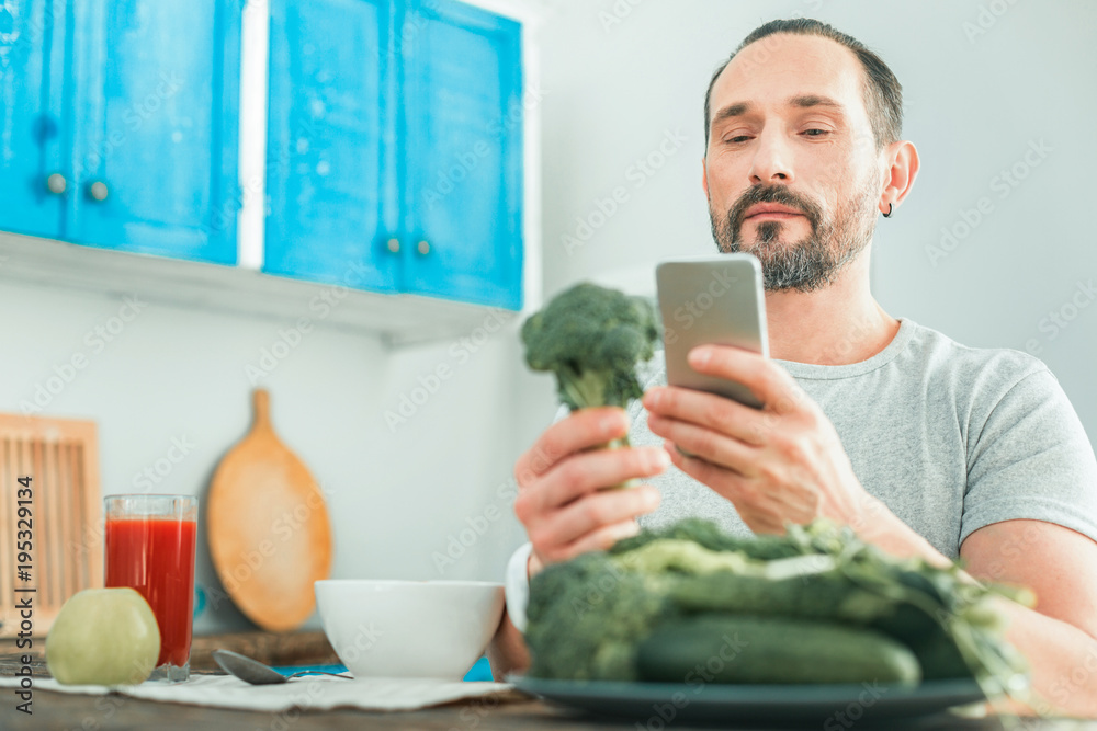 Fototapeta premium Check before eat this. Calm concentrated occupied man being in the kitchen looking at the cellphone holding a broccoli.
