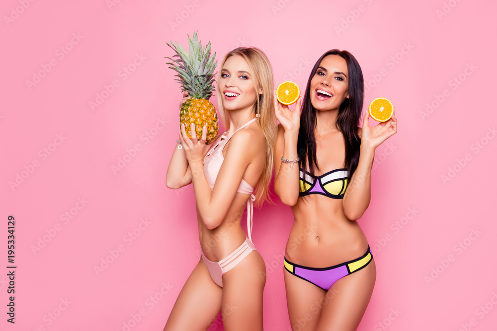Portrait of pretty, charming, laughing, stylish trendy ladies, tourists with modern hairstyle in swimsuit showing ananas and orange in hands, standing over pink background, brunette vs blonde - obrazy, fototapety, plakaty 