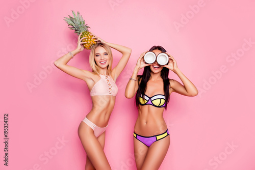 Portrait of hot, charming, stylish, pretty, trendy, crazy ladies, tourists in swim suits, blonde having ananas on her head, brunette having coconut on eyes' place, standing over pink background