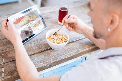 Nutritious treat. Skillful ambitious man eating healthy granola while taking spoon and studying article about biohacking