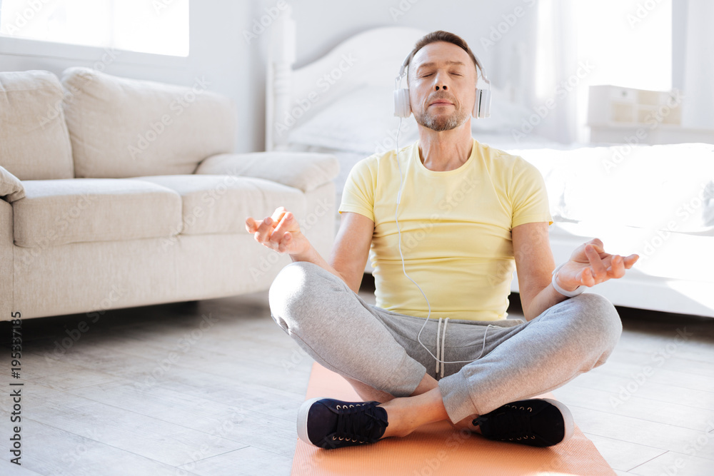 Right music. Charming handsome ambitious man meditating on mat board while sitting in lotus posture and controlling breath Right music. Charming handsome ambitious man meditating on mat board while sitting in lotus posture and controlling breath