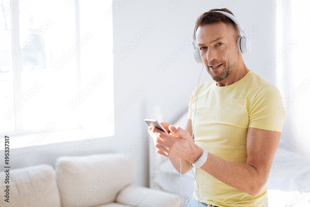 Modern gadget. Confident appealing smart man using device while looking at camera and wearing headphones Modern gadget. Confident appealing smart man using device while looking at camera and wearing headphones