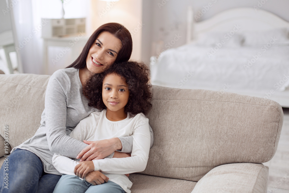 Happy together. Attractive alert young dark-haired woman smiling and hugging her daughter while sitting on the couch