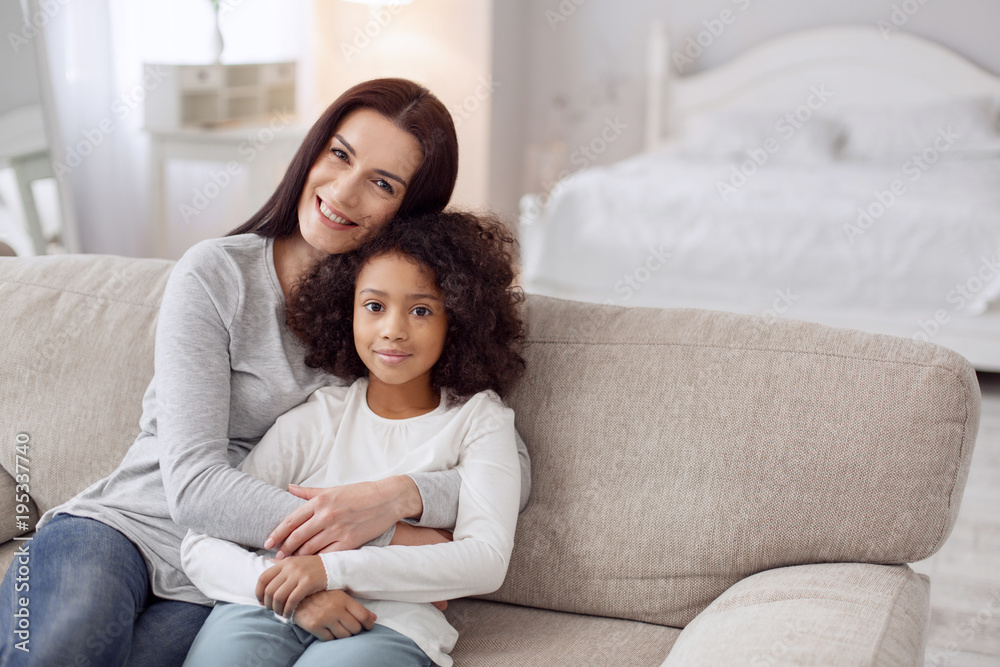 Happy together. Attractive alert young dark-haired woman smiling and hugging her daughter while sitting on the couch Happy together. Attractive alert young dark-haired woman smiling and hugging her daughter while sitting on the couch