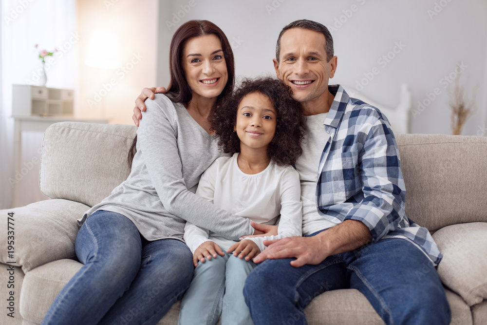 Happy family. Pretty joyful curly-haired girl smiling and sitting on the couch with her parents and they hugging her Happy family. Pretty joyful curly-haired girl smiling and sitting on the couch with her parents and they hugging her