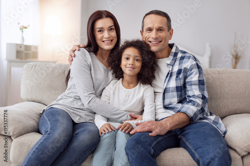 Happy family. Pretty joyful curly-haired girl smiling and sitting on the couch with her parents and they hugging her