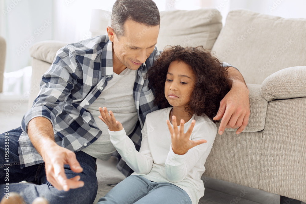Happy weekend. Handsome alert dark-haired man smiling and teaching his daughter to count on her fingers while sitting near her on the floor