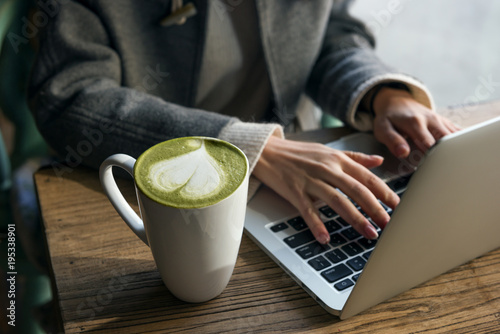 Beautiful young woman's hands busy working on her laptop sitting at wooden table in a coffee shop. And closeup of Matcha Latte.