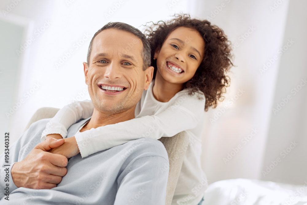 Beaming. Good-looking alert dark-haired father sitting in the arm-chair and smiling and his daughter standing behind him Beaming. Good-looking alert dark-haired father sitting in the arm-chair and smiling and his daughter standing behind him