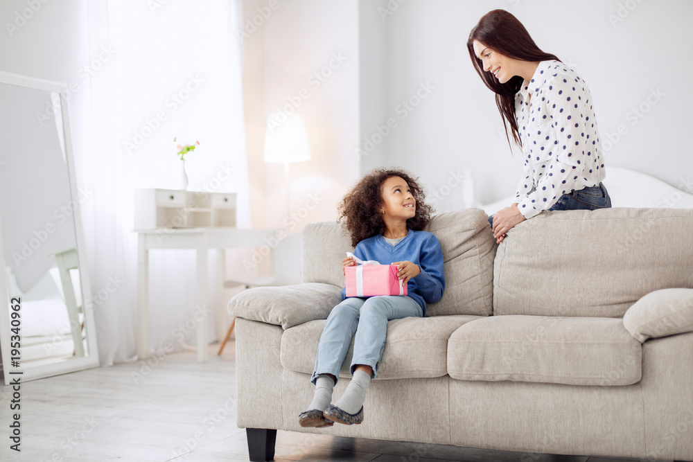 Happy celebration. Attractive cheerful curly-haired girl smiling and holding her gift while looking at her mother standing behind her