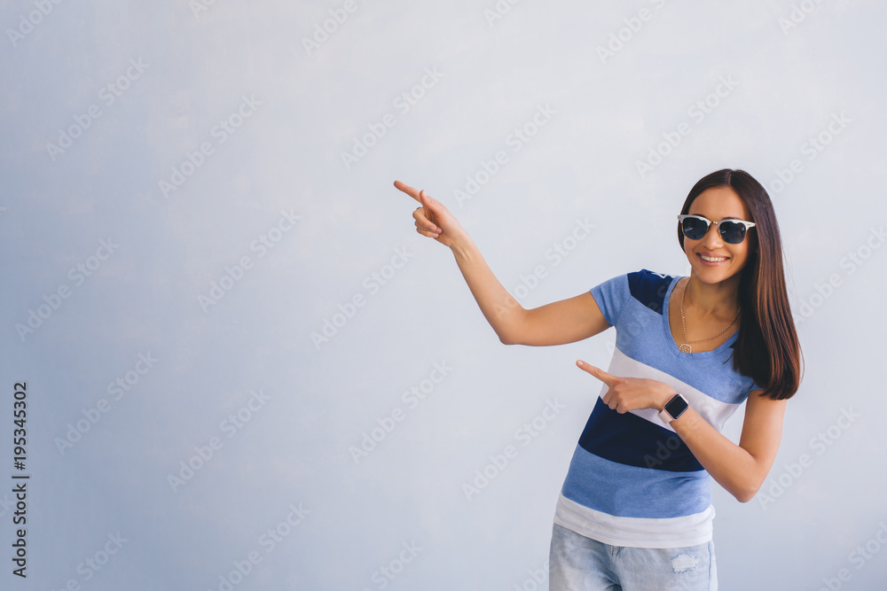 Look over there. modern youth woman, wearing casual clothes and sunglasses pointing copy space, smiling while standing against grey background