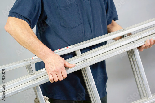 An electrician holds an aluminum ladder. The builder carries an aluminum stepladder on gray background. Worker of the plant with aluminum rung ladder photo