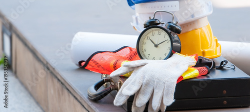 Stack of engineer safety helmets with construction area blueprint, black alarm clock with cotton gloves lay on nearly working area.