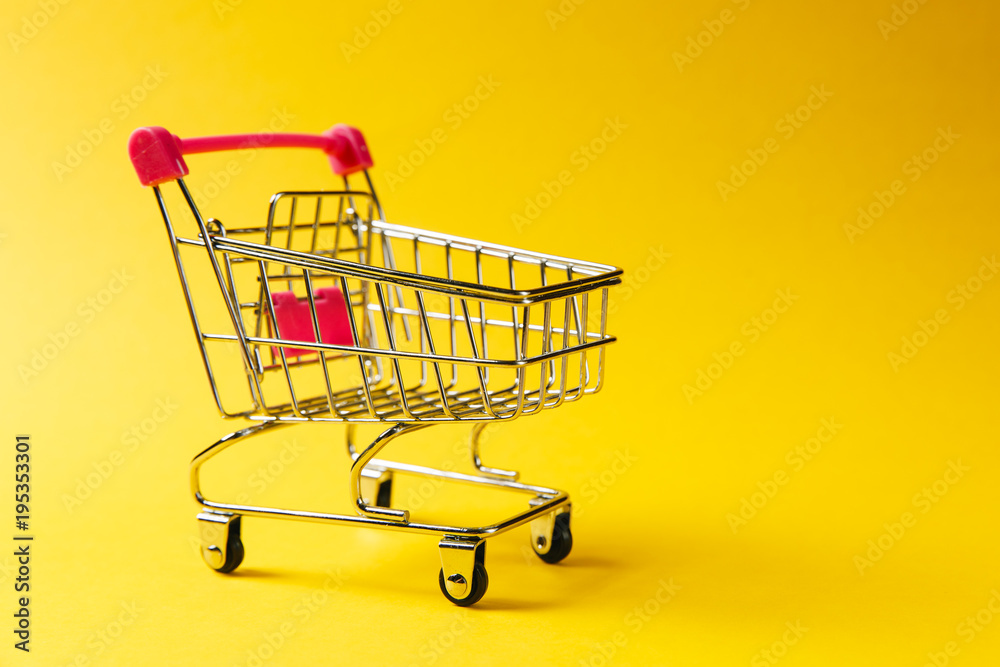 Close up of supermarket grocery push cart for shopping with black wheels and red plastic elements on handle isolated on yellow background. Concept of shopping. Copy space for advertisement Close up of supermarket grocery push cart for shopping with black wheels and red plastic elements on handle isolated on yellow background. Concept of shopping. Copy space for advertisement