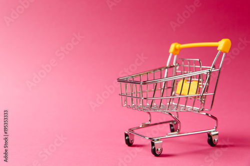 Close up of supermarket grocery push cart for shopping with black wheels and yellow plastic elements on handle isolated on pink background. Concept of shopping. Copy space for advertisement