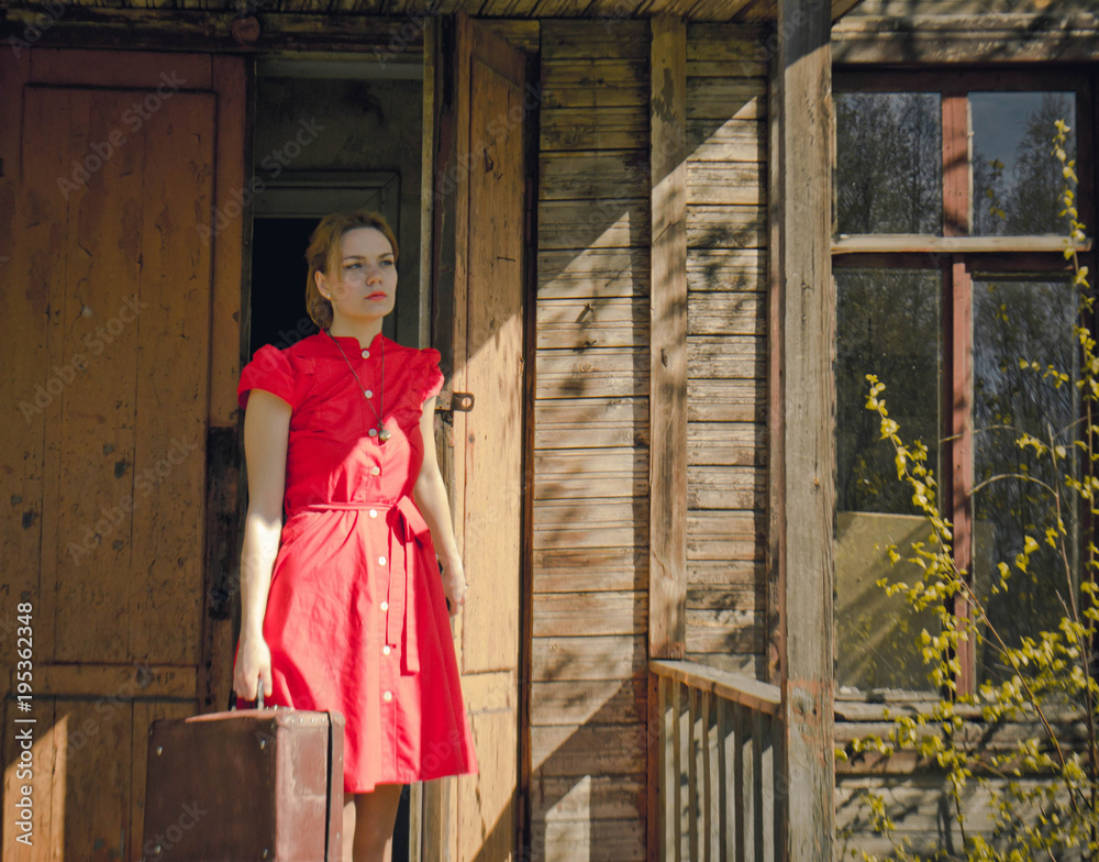 near the old wooden house girl in a red dress with a suitcase