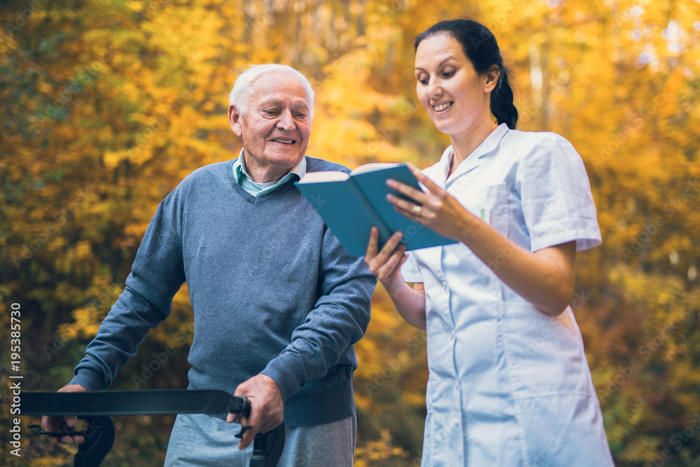 Smiling nurse reading book to senior man that uses walker with caregiver outdoor Smiling nurse reading book to senior man that uses walker with caregiver outdoor