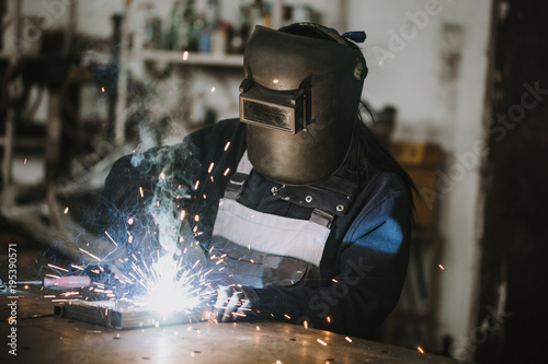 Strong and worthy woman welder doing hard job in car and motorcycle repair shop. She using welding machine to fix some metal bike parts.