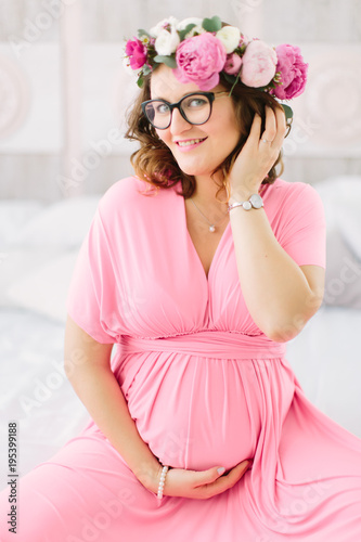 Young beautiful pregnant girl in a pink dress is ssitting on the bed with pregnant belly in her hand photo