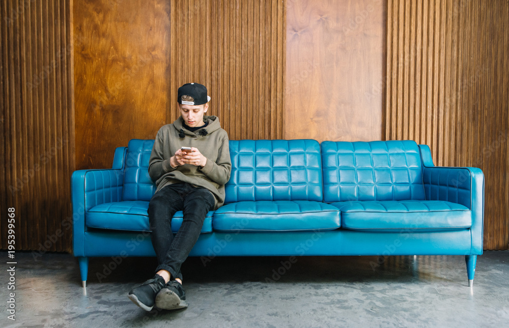 Fotografie Young man sitting on a blue couch and using a smartphone