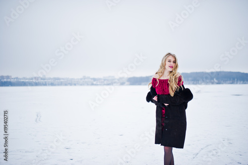 Elegance blonde girl in fur coat and red evening dress posed at winter snowy day.