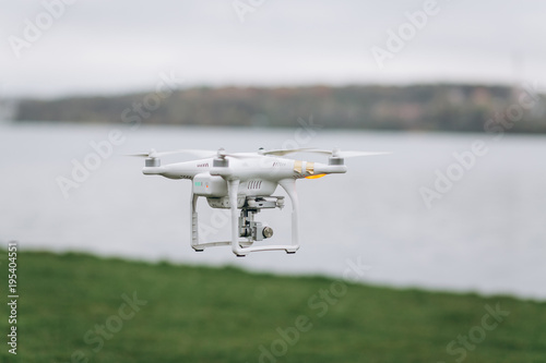 White quadrocopter is flying high in the air, taking photos, recording footage from above. Drone with four motors and propellers, camera and red warning lights.