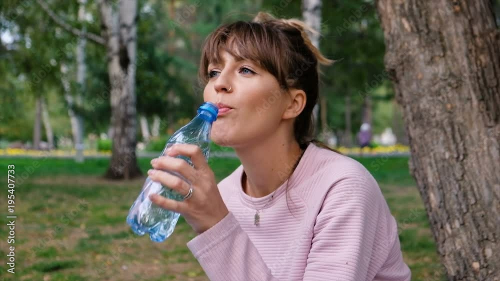 Attractive girl blonde drink water with pleasure outdoors. A young woman is drinking clean water from a plastic bottle. Hipster student drinks water from a bottle in the park and looking at the camera Attractive girl blonde drink water with pleasure outdoors. A young woman is drinking clean water from a plastic bottle. Hipster student drinks water from a bottle in the park and looking at the camera
